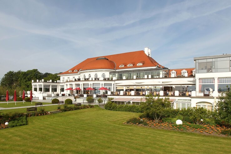 Blick auf das Gebäude und den Garten des Columbia Hotel in Travemünde Elegantes Hotel mit rotem Dach, großen Fenstern und Terrassen, umgeben von gepflegtem Garten mit Blumen und Rasen.