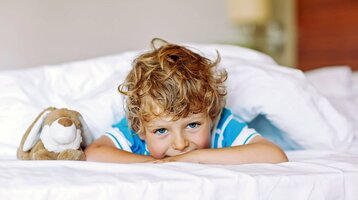 Kind liegt lächelnd auf einem Hotelbett mit Kuscheltier, umgeben von weißen Kissen. Foto: © Irina Schmidt - Fotolia.com