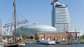 Exterior view of the ATLANTIC Hotel Sail City Bremerhaven Harbor view with sailing ship, modern glass building and historic brick building under a clear sky.