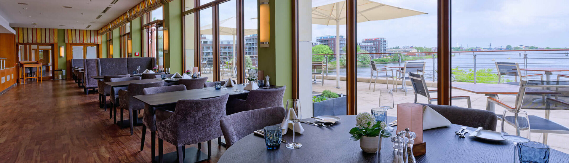Restaurant Harbour View im ATLANTIC Hotel Wilhelmshaven Elegantes Hotelrestaurant mit gedeckten Tischen, großen Fenstern und Blick auf eine Terrasse mit Fluss im Hintergrund.