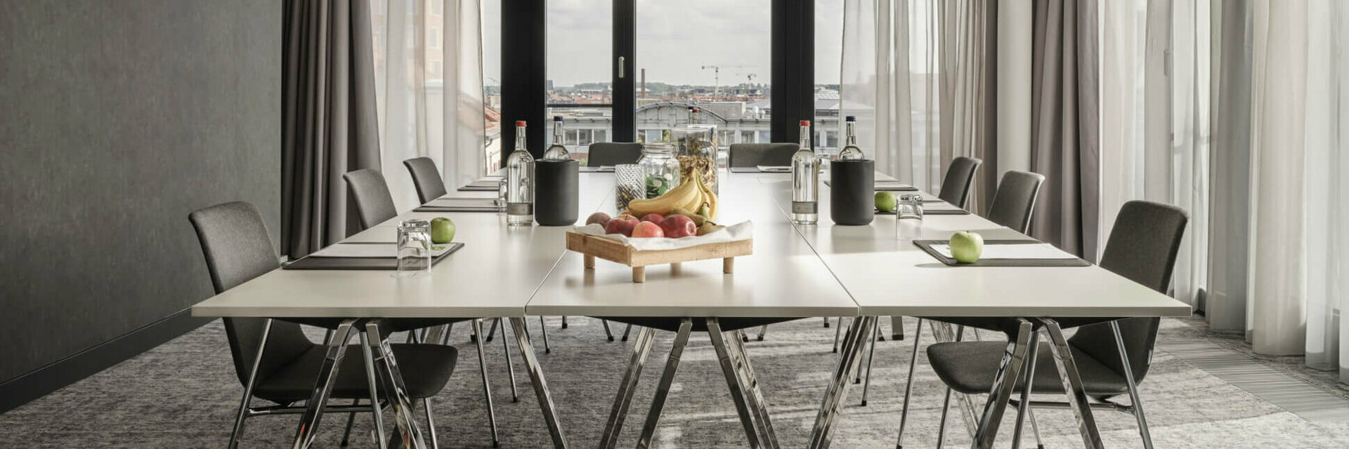 ATLANTIC Hotel Münster Tagungsangebot 15 % Moderner Konferenzraum im ATLANTIC Hotel Münster mit großem Tisch, Stühlen und einem Obsttablett, lichtdurchflutet durch Fenster.