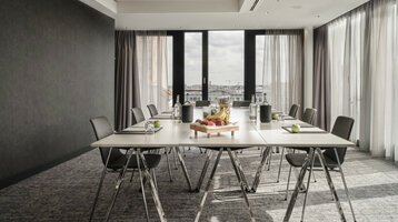 ATLANTIC Hotel Münster Tagungsangebot 15 % Moderner Konferenzraum im ATLANTIC Hotel Münster mit großem Tisch, Stühlen und einem Obsttablett, lichtdurchflutet durch Fenster.