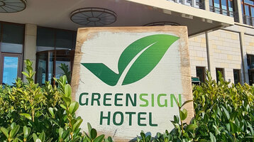 GreenSign Certificate | ATLANTIC Hotel Wilhelmshaven Facade of a modern hotel with "Greensign Hotel" sign and green plants in the foreground.