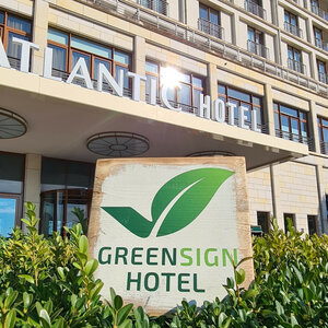 GreenSign Zertifikat | ATLANTIC Hotel Wilhelmshaven Fassade eines modernen Hotels mit "Greensign Hotel"-Schild und grünen Pflanzen im Vordergrund.