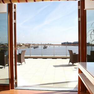 Ausblick auf den Hafen vom Restaurant Harbour View des ATLANTIC Hotel Wilhelmshaven Helle Terrasse mit Blick aufs Wasser, modernen Möbeln und Pflanzen im Vordergrund.
