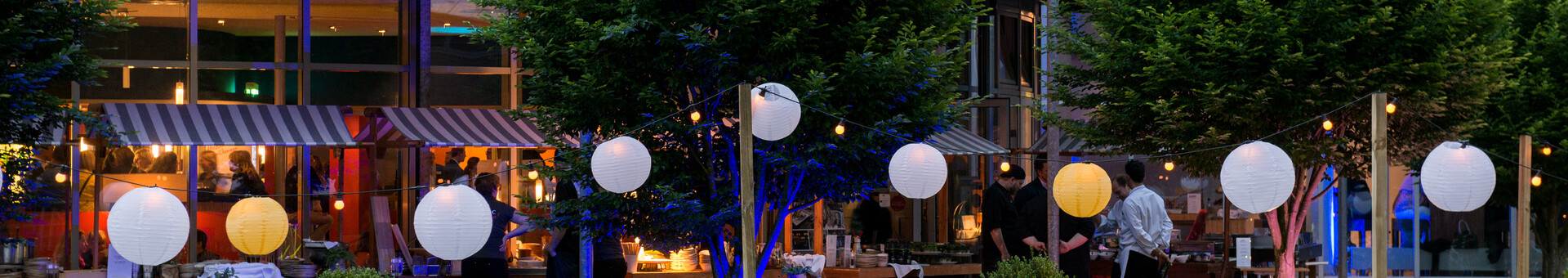 Outdoor area of the ATLANTIC Hotel Universum with illuminated lanterns, tables and chairs, surrounded by trees.