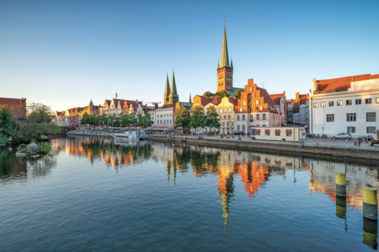 The shore of the Trave in Lübeck Historic buildings and church towers are reflected in the water of a river under a clear sky.