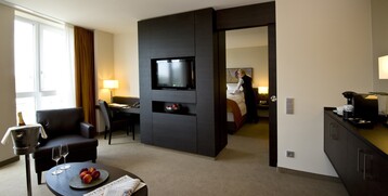 Interior view of a Suite in the ATLANTIC Grand Hotel in Bremen Modern hotel room with sitting area, desk, TV, bed in the background and view of a person making the bed.