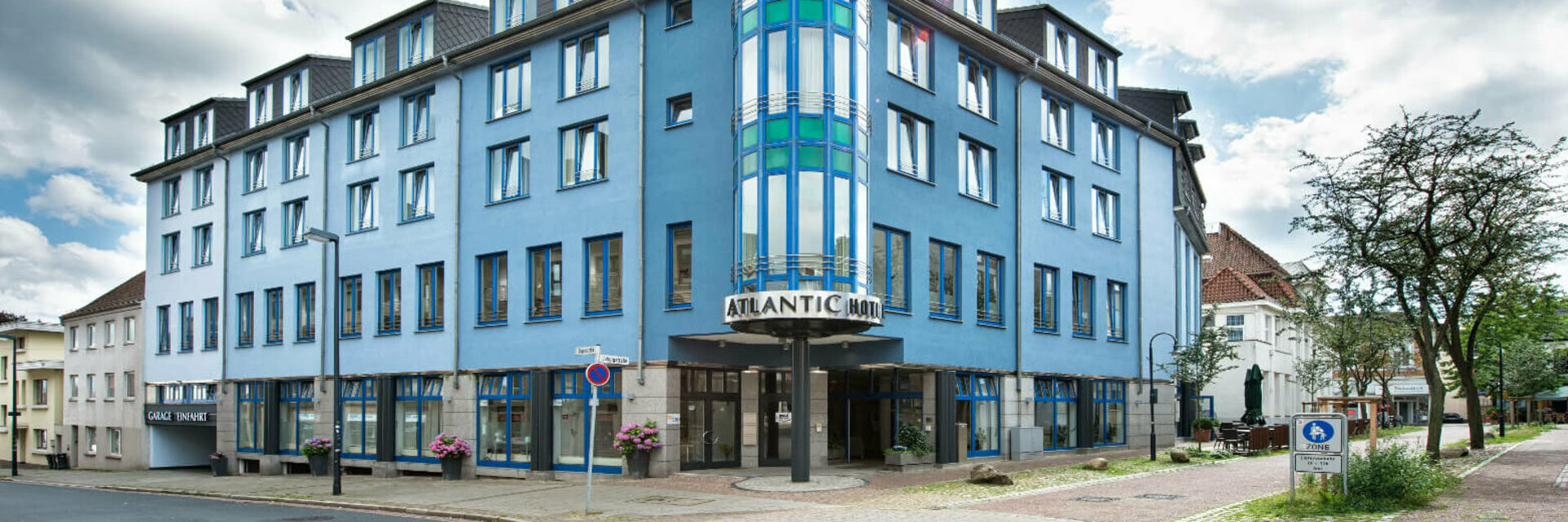 ATLANTIC Hotel Vegesack Bremen Blue building of the ATLANTIC Hotel Vegesack with striking tower and entrance, surrounded by trees and cobbled street.