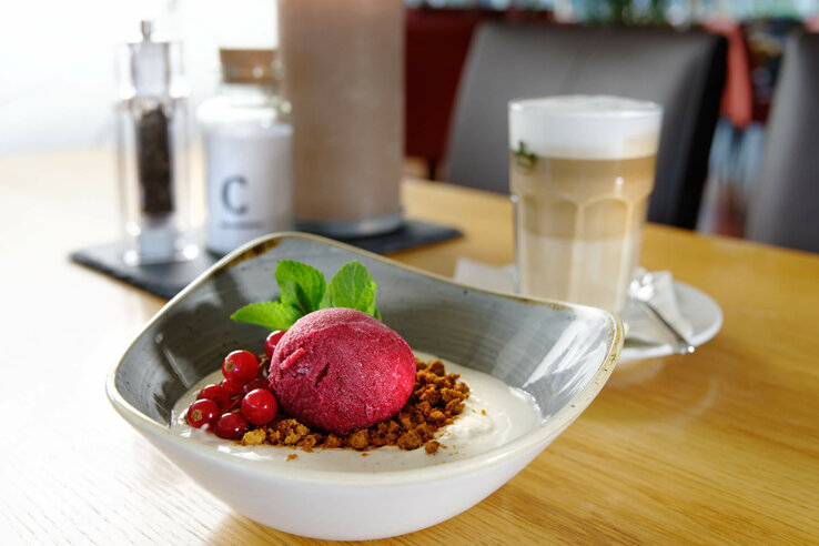 Food from the ATLANTIC Hotel Bremen Universum Red sorbet ball with berries and mint on dessert, next to it a latte macchiato on a wooden table in the ATLANTIC Hotel Universum.