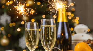 Zwei Sektgläser mit Wunderkerzen, Champagnerflasche und Orangen vor unscharfem, festlichem Hintergrund.