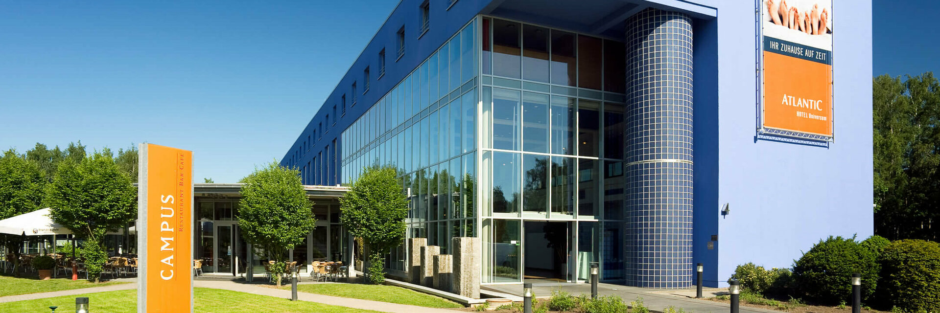 Außenansicht des ATLANTIC Hotel Universum ATLANTIC Hotel Universum in Bremen: Modernes, blaues Gebäude mit Glasfassade, umgeben von gepflegtem Rasen und Bäumen.
