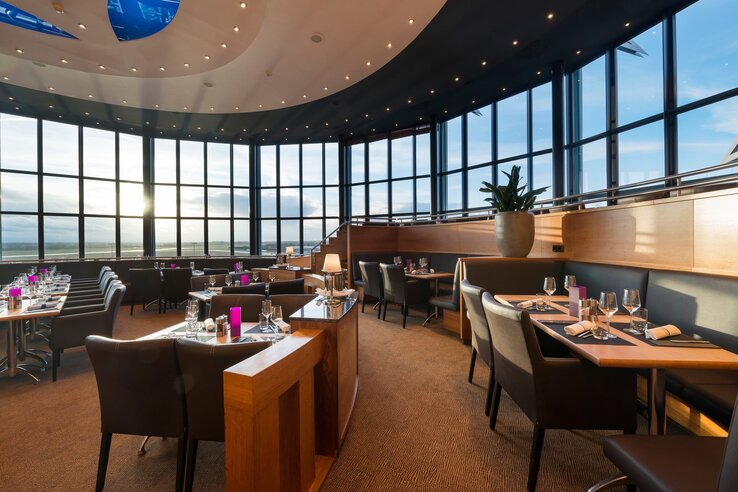 Restaurant Blixx im ATLANTIC Hotel Airport Modernes Restaurant mit großen Fenstern, elegant gedeckten Tischen und stimmungsvoller Beleuchtung bei Sonnenuntergang.