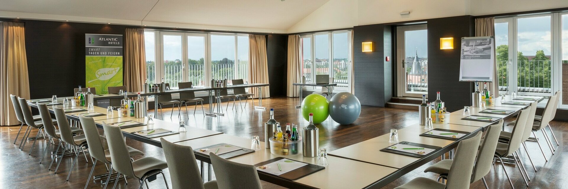 Roof Lounge im ATLANTIC Hotel Lübeck Moderner Konferenzraum mit U-förmiger Tischanordnung, Stühlen, Flipchart und großen Fenstern mit Blick ins Grüne.