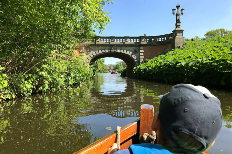 The Bürgerpark in Bremen A child in a boat on a river passing under a stone bridge with lanterns, surrounded by lush greenery.