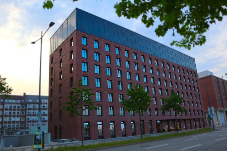 Unique Kiel by ATLANTIC Hotels Modernes Hotelgebäude aus rotem Backstein mit großen Fenstern, umgeben von Bäumen und Straße im Vordergrund.