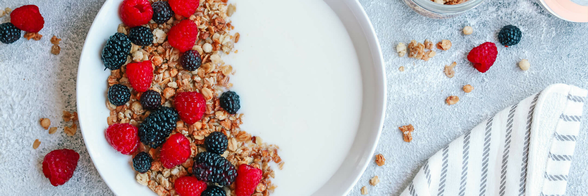 Frühstück in ATLANTIC Hotel Wilhelmshaven Schale mit Joghurt, Müsli, Himbeeren und Brombeeren, umgeben von gefüllten Gläsern und einem gestreiften Tuch.