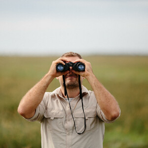 Man with binoculars | ATLANTIC Hotel Wilhelmshaven Man in beige shirt observes nature with binoculars in a wide, green landscape.