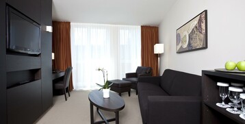 Interior view of the seating and Tv area of the ATLANTIC Grand Hotel in Bremen Modern hotel room with black sofa, coffee table, TV, desk, artwork on the wall and large windows.