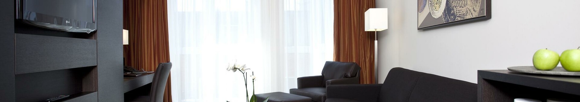 Interior view of the seating and Tv area of the ATLANTIC Grand Hotel in Bremen Modern hotel room with black sofa, coffee table, TV, desk, artwork on the wall and large windows.