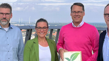 GreenSign certified | ATLANTIC Hotels Four people on a viewing platform, one holding a Greensign hotel sign, wind turbines and city view in the background.