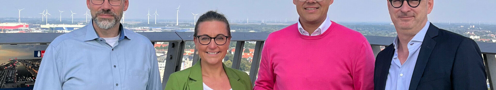 GreenSign certified | ATLANTIC Hotels Four people on a viewing platform, one holding a Greensign hotel sign, wind turbines and city view in the background.