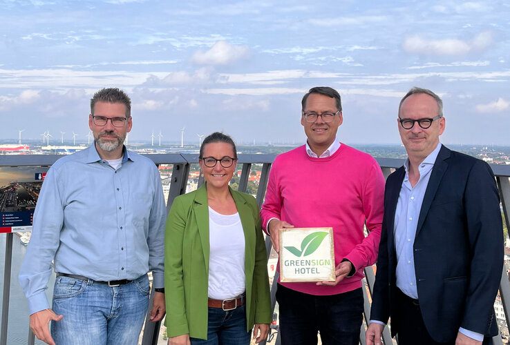 GreenSign certified | ATLANTIC Hotels Four people on a viewing platform, one holding a Greensign hotel sign, wind turbines and city view in the background.