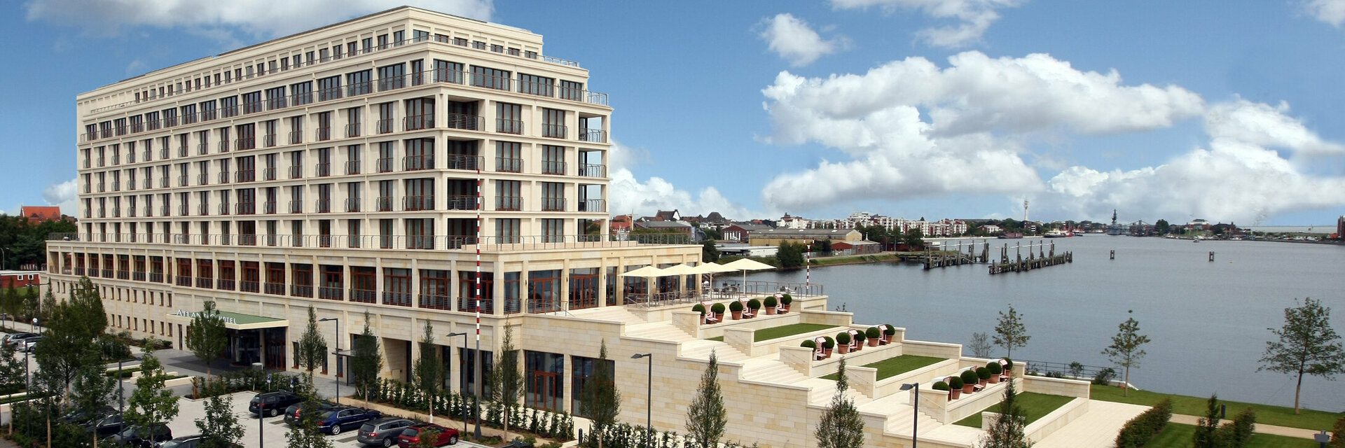 exterior view of the ATLANTIC Hotel Wilhelmshaven Modern hotel on the waterfront with large windows, terraces and well-tended garden, surrounded by blue sky and clouds.
