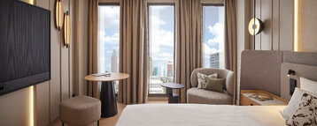 Modern hotel room at the ATLANTIC Hotel Frankfurt with a large bed, seating area with armchair and small table, and floor-to-ceiling windows offering views of the city skyline.