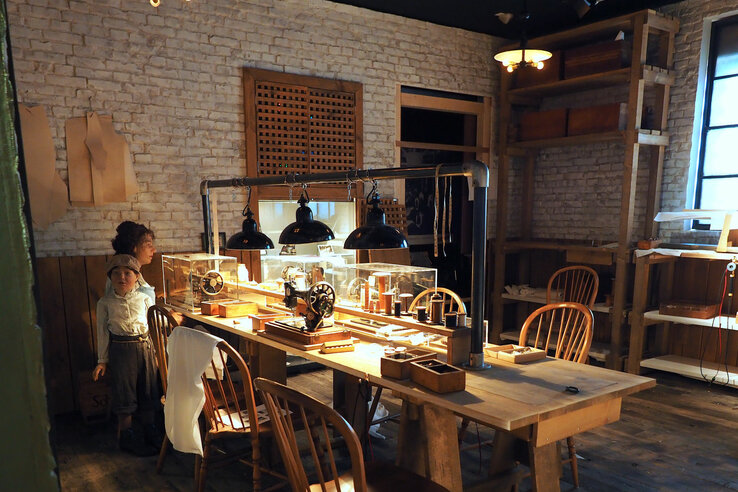 Cozy workshop with wooden table, sewing machines and warm light, two people sitting at a window.