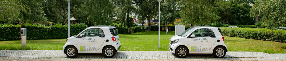 Electric filling stations | Service of the ATLANTIC Hotels Two white electric Smarts with the "Atlantic Hotels" logo are parked next to each other on a paved path, surrounded by trees.