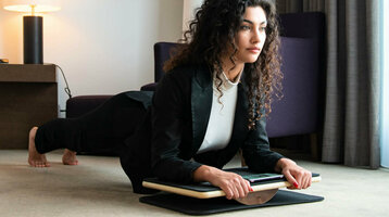 Plank pads available in selected ATLANTIC Hotels Woman in business attire doing plank exercise on balance board in modern hotel room.