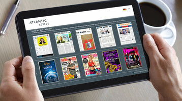 ATLANTIC Hotels | Mediabox Tablet shows digital magazine selection in a hotel, held by hands, next to a cup of coffee and a pen.