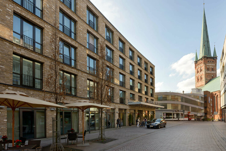 ATLANTIC Hotel Lübeck – Außenansicht Fassade des ATLANTIC Hotel Lübeck mit modernen Fenstern, Sonnenschirmen und einem Kirchturm im Hintergrund.