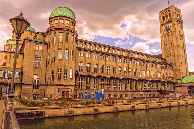 German Museum Munich Historic building with green domed roof and tower on the riverbank under a cloudy sky.
