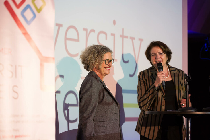 Claudia Kessler - Diversity Preis Gewinnerin 2016 Zwei Frauen auf einer Bühne, eine spricht in ein Mikrofon, im Hintergrund ein buntes Logo und der Schriftzug "Diversity".