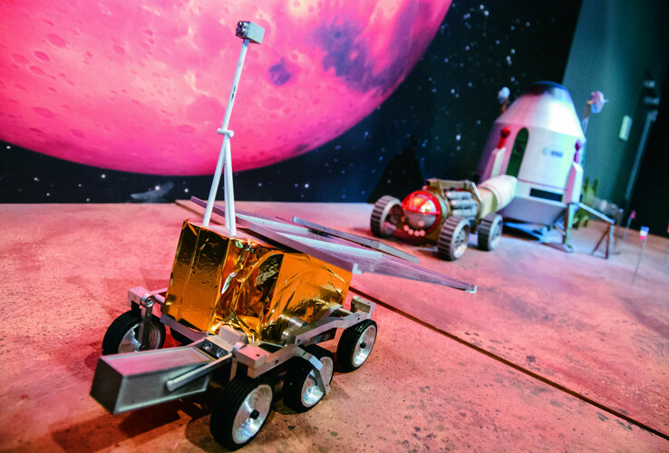 guided tours Model of a lunar rover and a space capsule in front of a large, pink-colored planetary background.