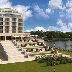 Modernes Hotel am Fluss mit Terrassen, Liegestühlen und Sonnenschirmen, umgeben von grüner Landschaft und blauem Himmel.