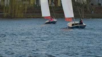 Angebot Segeln in Münster l ATLANTIC Hotel Münster Zwei Segelboote auf einem See vor einer Baumreihe und Häusern, mit ATLANTIC Hotel Münster Logo auf den Segeln.
