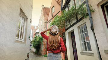 Frau in roter Bluse und Hut spaziert durch enge, malerische Altstadtgasse mit Fachwerkhäusern und Kopfsteinpflaster.