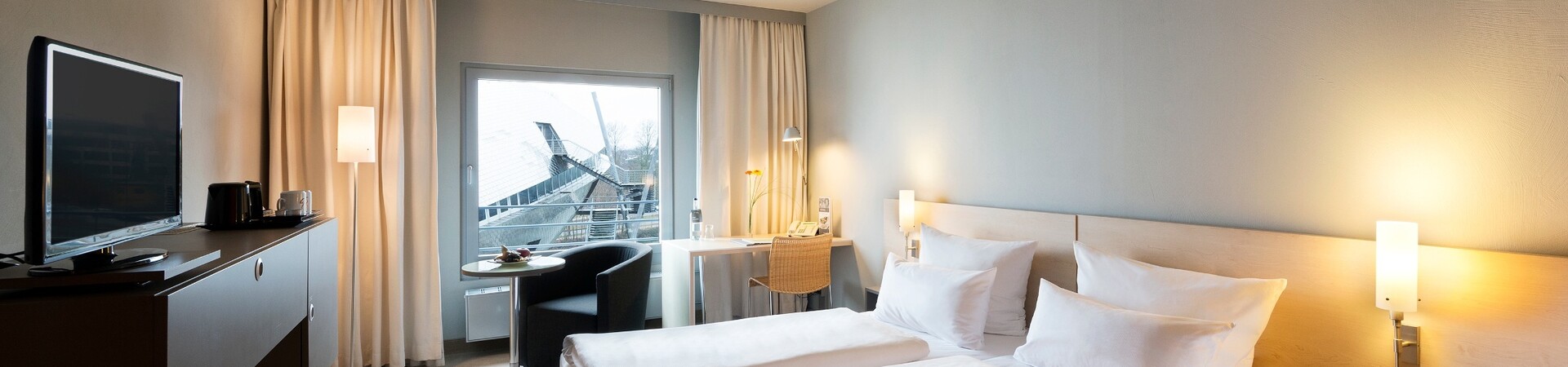 Comfort Room in the ATLANTIC Hotel Universum Bremen Bright hotel room with double bed, desk, TV and view of modern architecture through large window.