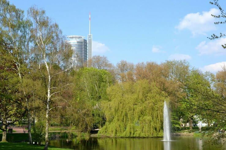 Park in the city of Essen High-rise building behind green park with pond and bubbling fountain, blue sky in the background.