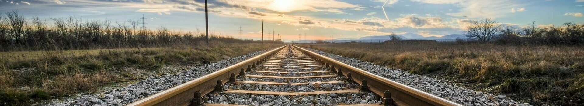 Travelling by rail to the ATLANTIC Hotels Tracks lead into the horizon, surrounded by fields, under a dramatic sunset sky.