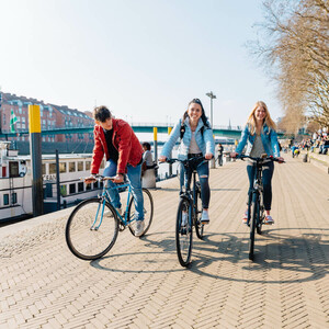 Bike paths in Bremen and surroundings l Bike it Schlachte ©WFB - Jonas Ginter Three people riding bicycles along a waterfront promenade in sunny weather, with boats and buildings in the background.