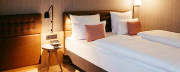 Modernes Hotelzimmer mit großem Doppelbett, weißer Bettwäsche und mehreren Zierkissen. Rechts und links am Kopfteil befinden sich Wandlampen, daneben steht ein kleiner runder Holztisch mit Telefon und Notizblock.