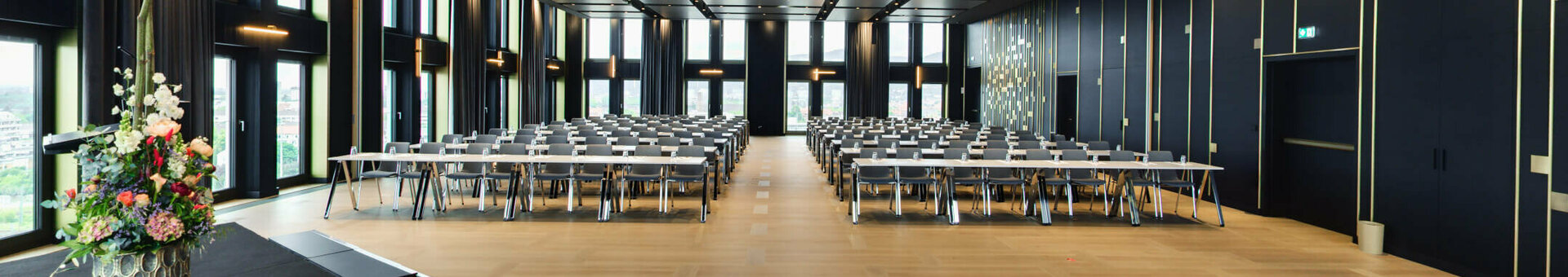 ATLANTIC Hotel Heidelberg Ballsaal parlamentarisch Modern conference room with large windows, rows of tables and chairs, flower arrangement on stage.