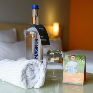 Welcome set in the hotel room: water bottle, glass, towel and jogging map on a table in front of the bed.