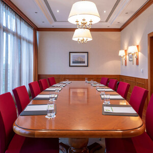 Conference room Münster in the ATLANTIC Hotel Wilhelmshaven Elegant conference room with large wooden table, red chairs, notepads and glasses of water, illuminated by large windows.