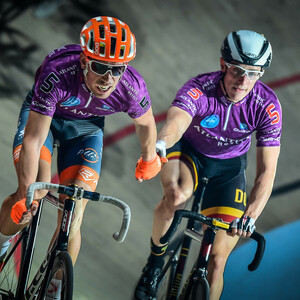 Two cyclists in purple jerseys ride side by side on a track and shake hands.