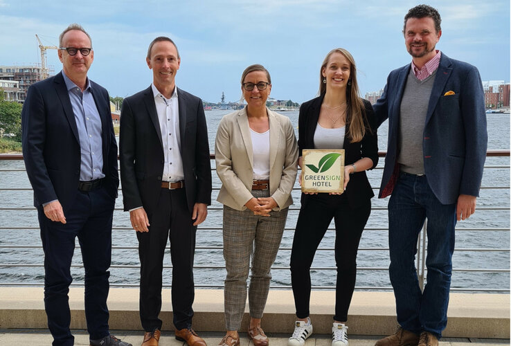 GreenSign Certificate| ATLANTIC Hotels Five people stand smiling on a riverbank, one holding a "Greensign Hotel" sign.